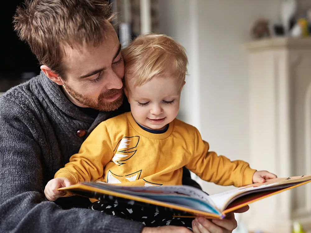 An image of a father and his child, the father with a serious expression on his face and the child looking up at him with a concerned expression. The background is blurred and there are no other visible details in the image.