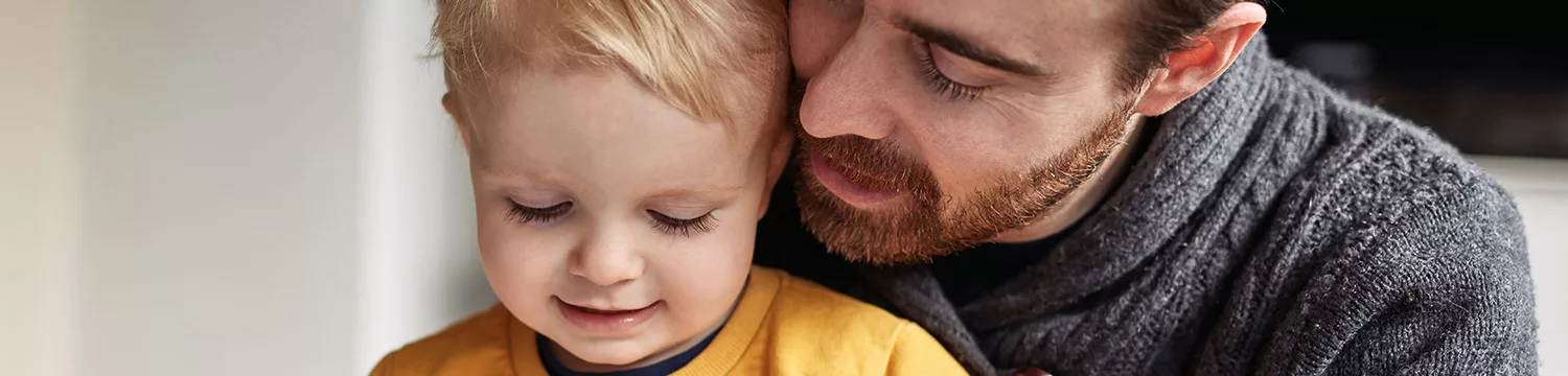 An image of a father and his child, the father with a serious expression on his face and the child looking up at him with a concerned expression. The background is blurred and there are no other visible details in the image.